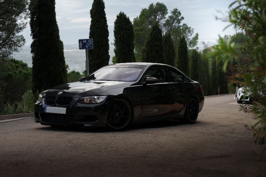 Black BMW 335i E92 parked on a coastal road with cypress trees
